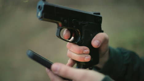 a close-up view of a man in a green jacket holding a cell phone and a gun, with his finger on the trigger, using one of his finger to operate his phone