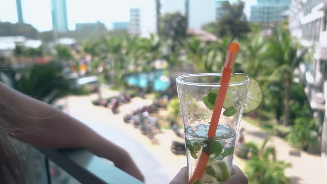 소녀는 흐릿한 리조트 마당을 반대하는 호텔 발코니에 서 있습니다.