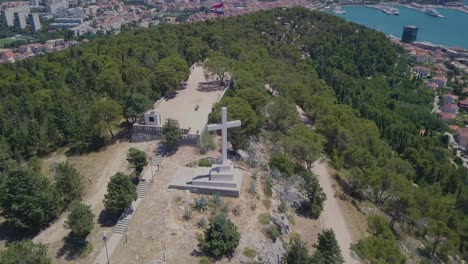 flying around the white stone cross, keeping it as the main subject with the port and city of split in the background