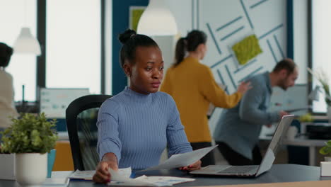 African-american-startup-employee-holding-business-papers-with-charts-looking-at-laptop-screen