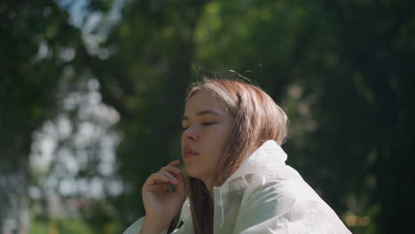 young woman removes raincoat hood, resting her hand on her chin, lost in thought, the soft sunlight illuminates her pensive expression against a gently blurred green backdrop