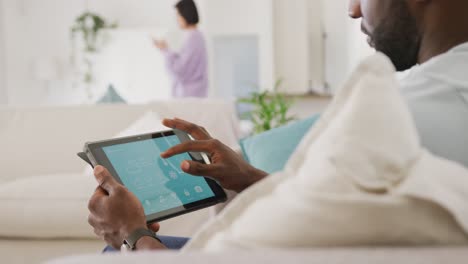 diverse couple sitting on couch, using tablet and walking in living room