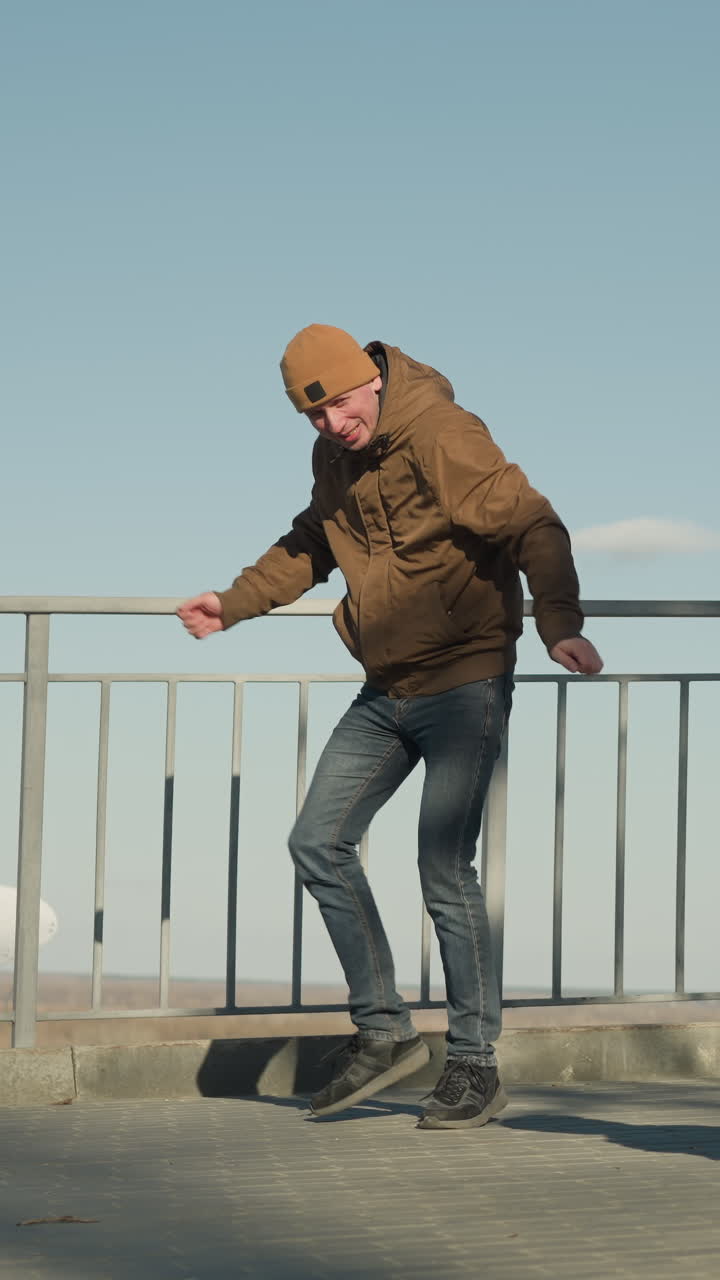 Close up of father and son dancing close to an iron rail on a walkway, the father wears a brown jacket and jeans while the son wears a black shinny jacket and jeans