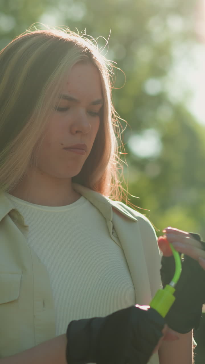 dama al aire libre con guantes negros examina la boquilla de la bomba verde de cerca bajo la luz del sol, su enfoque es intenso, resaltado por el fondo natural de la vegetación exuberante y los árboles iluminados por el sol
