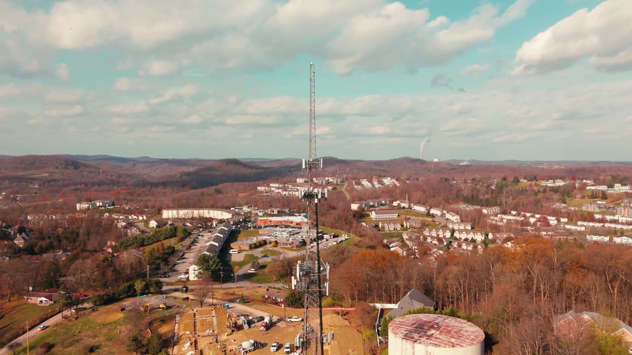 Orbita aerea attorno a una torre di telecomunicazioni sopra case suburbane circondate da alberi spogli sotto un cielo autunnale parzialmente nuvoloso.
