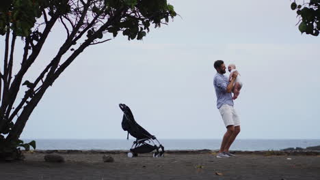 Inmitten-Von-Meer-Und-Strand-Fängt-Ein-Porträt-Den-Zärtlichen-Kuss-Eines-Vaters-Für-Sein-Baby-Ein.-Die-Urlaubsfamilie-Strahlt-Freude-Aus,-Papa-Hält-Das-Kleinkind-Liebevoll