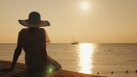 Una-Mujer-Joven-Se-Encuentra-Con-El-Amanecer-En-El-Muelle,-Se-Sienta-Y-Mira-El-Sol-Y-El-Barco-En-El-Mar-Dre