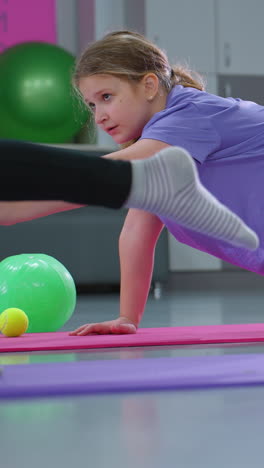 mano borrosa en primer plano con niños involucrados en una sesión de entrenamiento de equilibrio, una chica con una camisa púrpura sostiene una postura con un bloque rosa en la espalda, mientras otro niño se estira en una alfombra de yoga