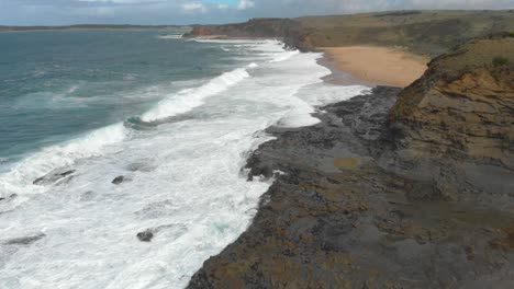 aerial shot moving forward from high up looking over coastal cliffs and large waves crashing onto the beach and rock shelf