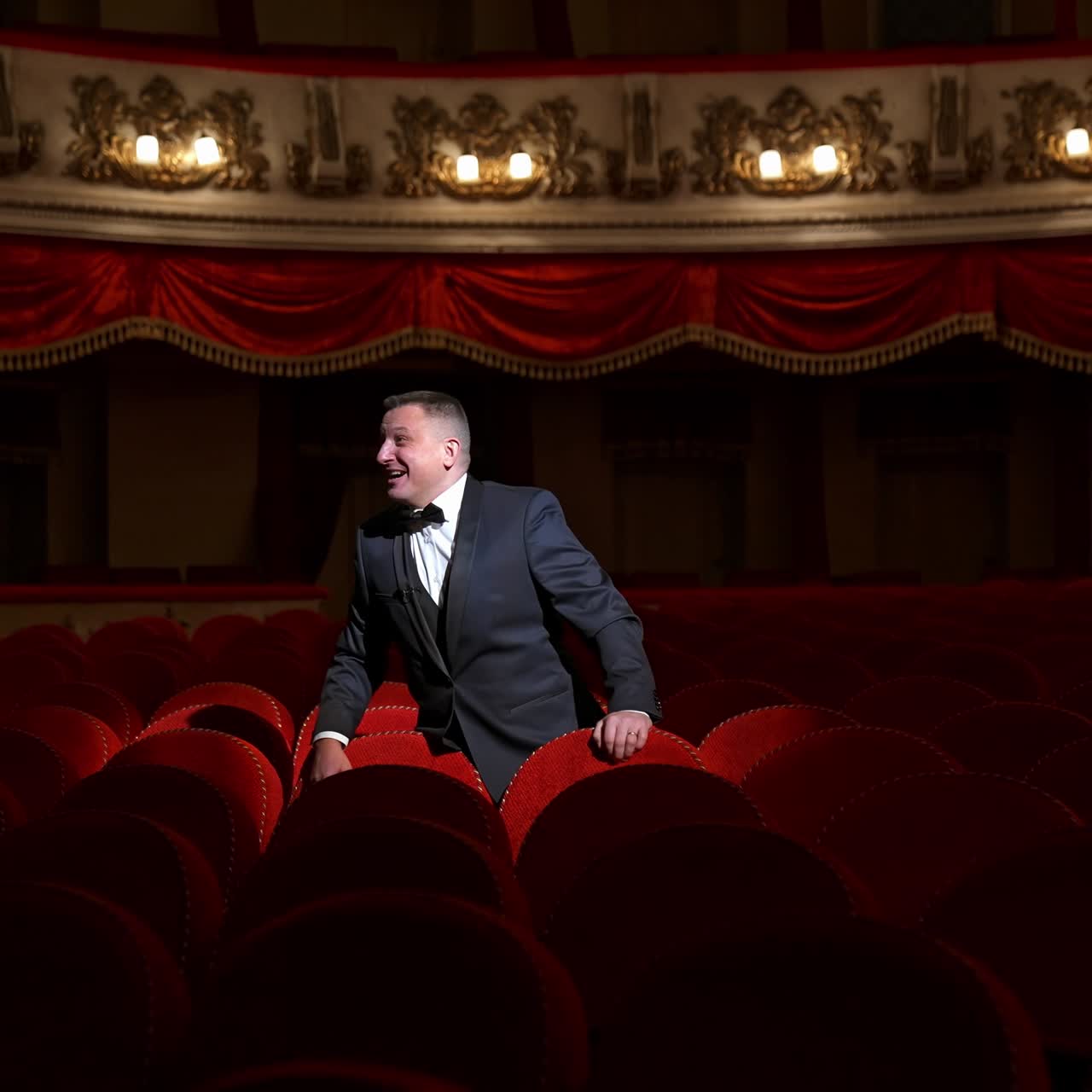 Man stands alone among velevet chair in classic theater. Male in classic suit invites public come in with gesture. Time before performance