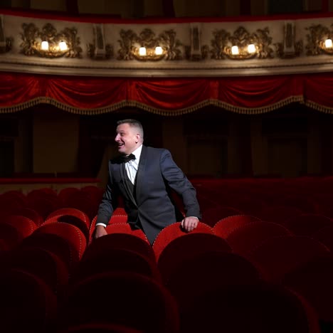 man stands alone among velevet chair in classic theater. male in classic suit invites public come in with gesture. time before performance