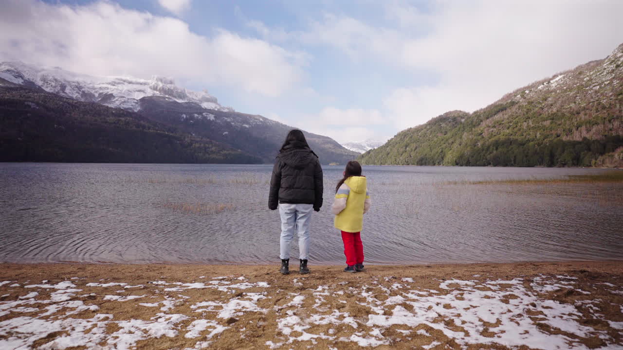 A woman and child walk toward the calm shores of Lago Falkner in Neuquén, Argentina, with snow-dusted sand, alpine forest, and Andean peaks in the background