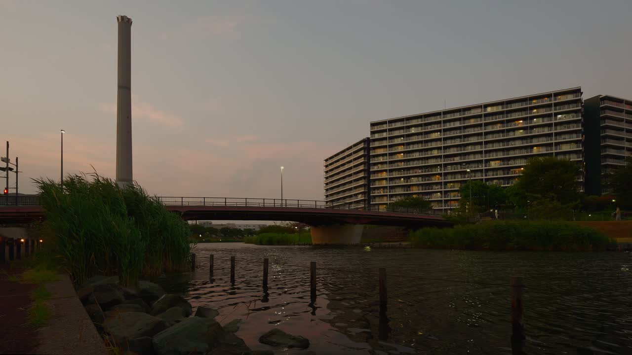 A wide shot of a tranquil river with a bridge, a tall smokestack, and a large apartment building