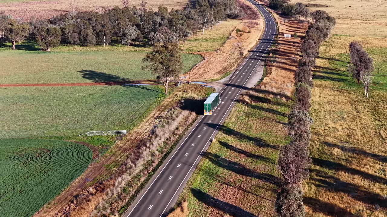 Aerial view of a green truck traveling on a paved road through open farmland, with long morning shadows and clear, natural lighting
