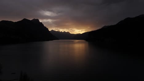 drone footage slowly pulls back, stunning sunrise hidden behind dark mountains with walensee lake beautifully centered beneath cloudy sky at wessen, switzerland.