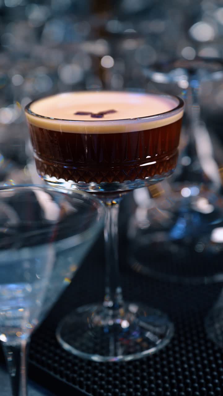 Cocktail espresso martini scuro in un bicchiere coupe di cristallo sfaccettato. Un cocktail marrone con strato di schiuma e guarnizione, presentato in un bicchiere dalla superficie lavorata su un bancone da bar scuro.