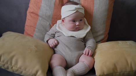 adorable baby girl sitting alone on sofa supported with cushions wearing a bib and headband