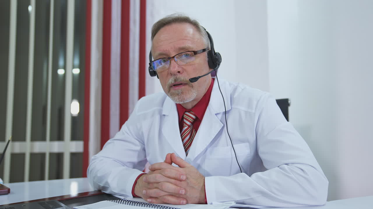 Serious doctor giving audio consultation call. Professional senior therapist sitting in medical office with headset and talking. Remote consultation.