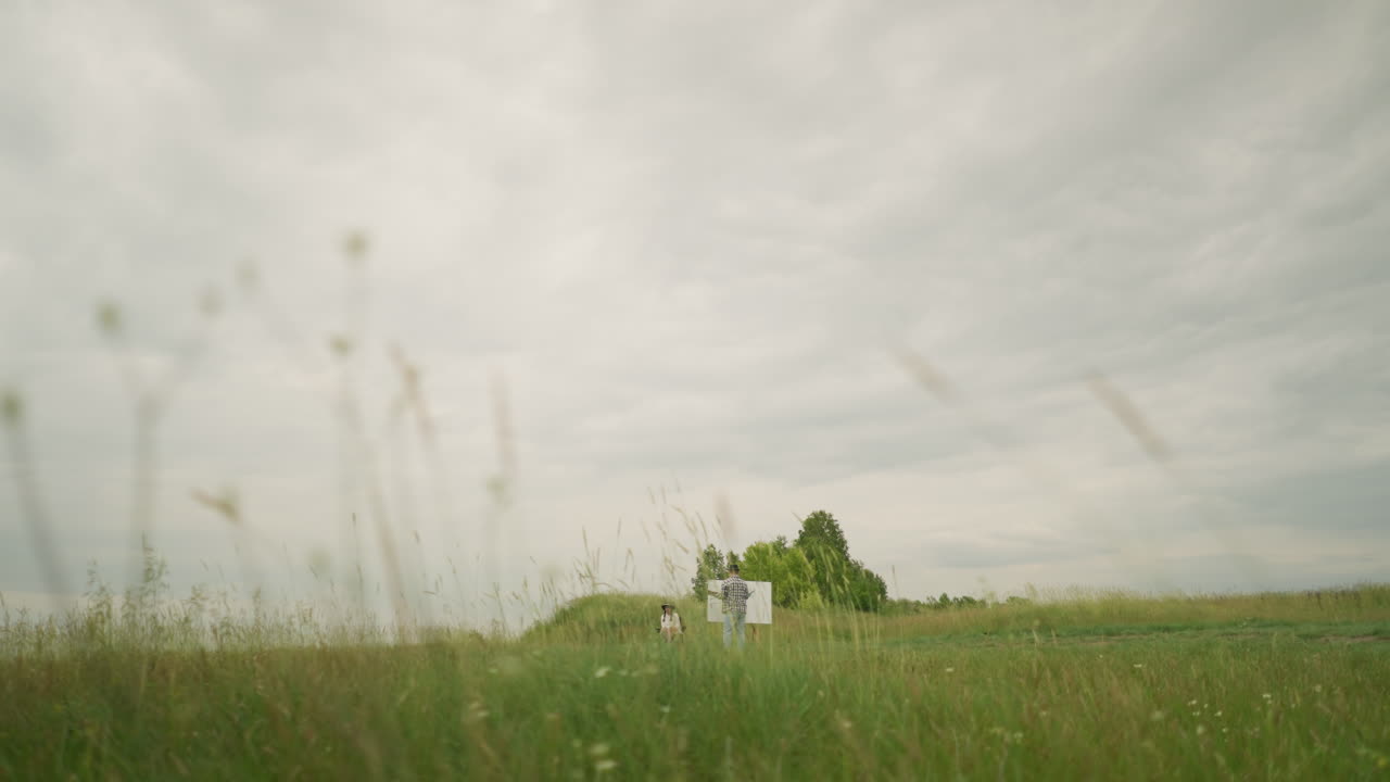 모자와 체크색 셔츠를 입은 화가의 뒷면은 구름이 은 하늘 아래 울창한 잔디 한가운데의 보드 위에 그림을 그리는 데 초점을 맞추고 있습니다.