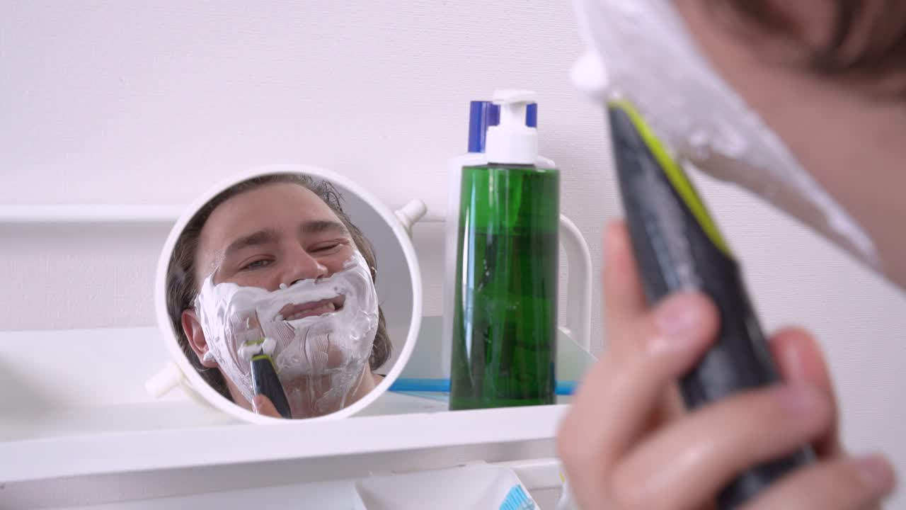 reflejo de un hombre con espuma de afeitar en la cara en un pequeño espejo redondo. un tipo se afeita el rastro y el bigote con una navaja eléctrica. cepillo de dientes y producto cosmético en el estante del baño. tratamientos diarios por la mañana