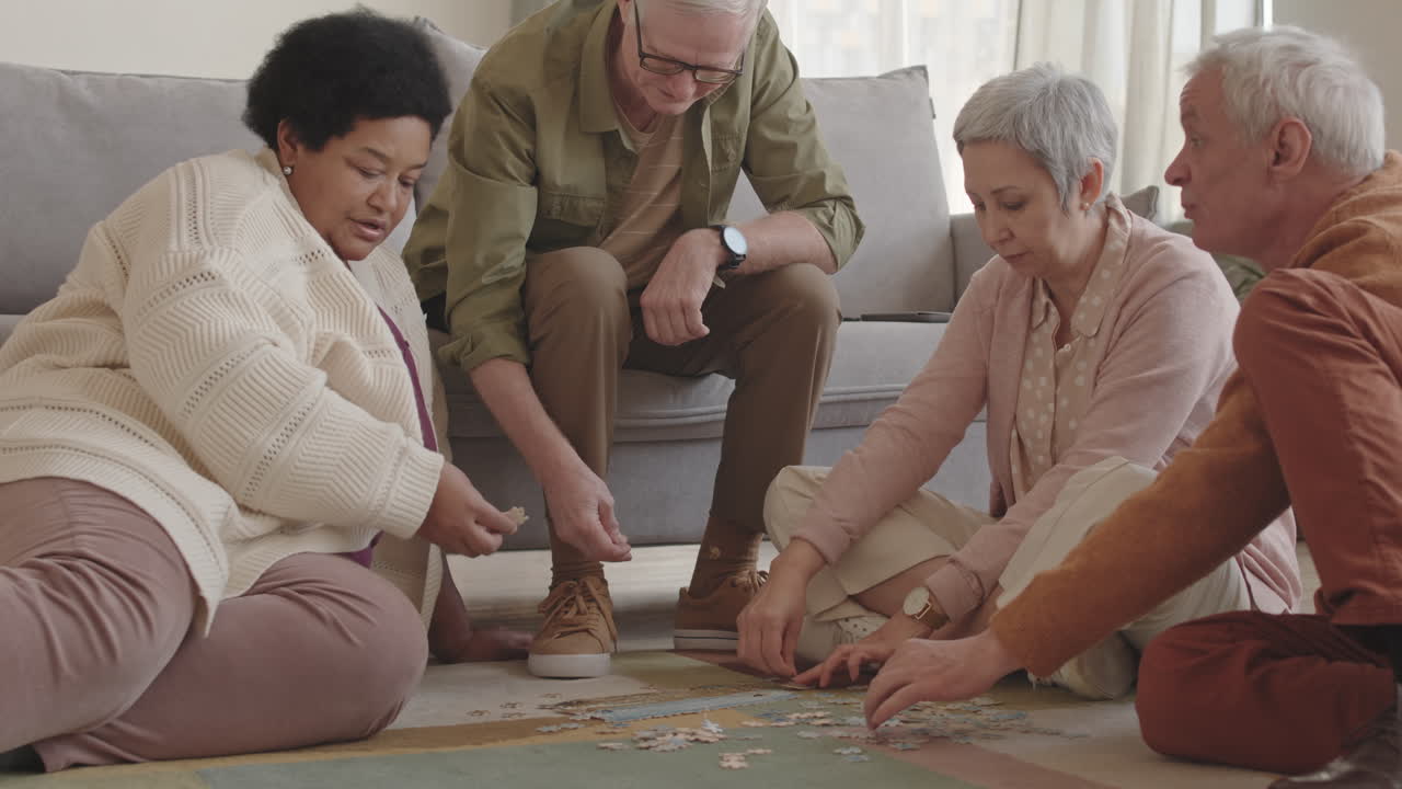 Medium long shot of company of diverse senior friends sitting on floor, talking, smiling, assembling puzzle