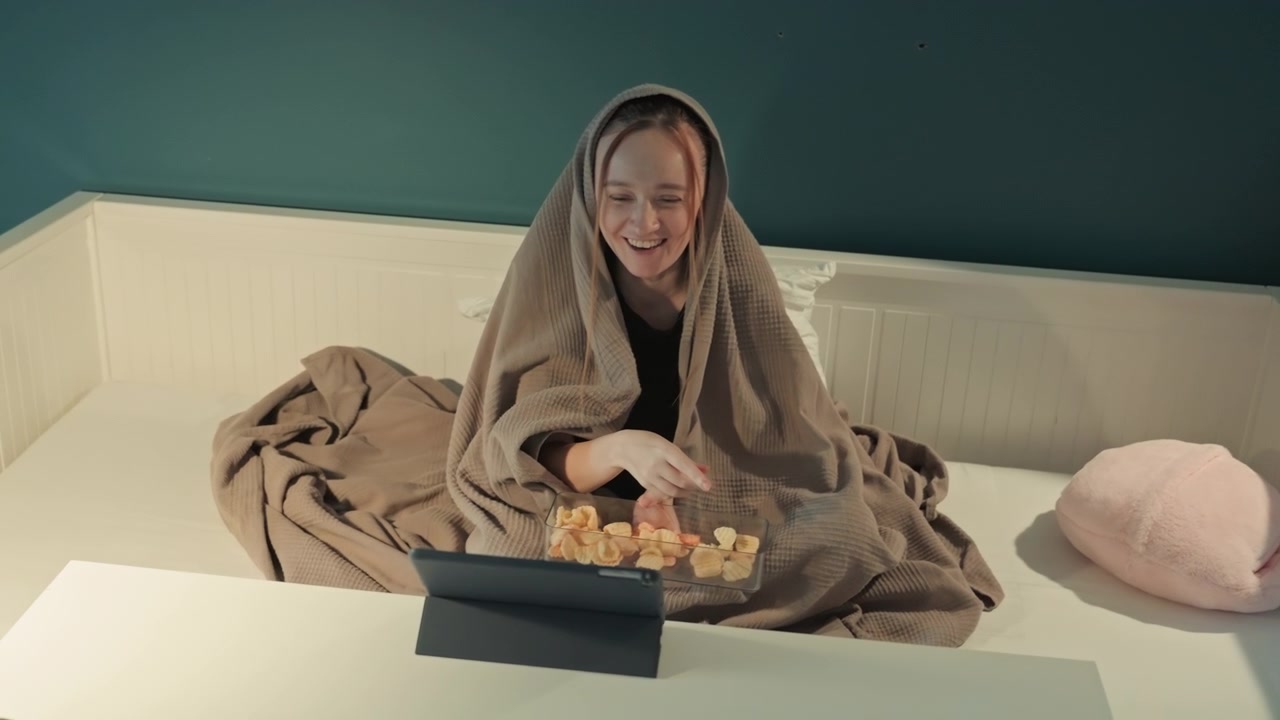 Giovane donna seduta sul letto avvolta in una coperta marrone che si allunga. Ambiente della camera da letto con parete color verde acqua, boiserie bianca e cuscino rosa peluche. Primo piano di una scena interna rilassata che mostra una selezione di snack.