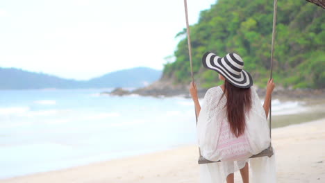 back of young sexy asian woman with big striped hat swings on seafront swing relaxing on tropical island