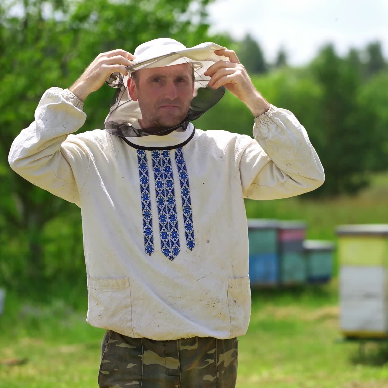 Portrait of beekeeper on apiary. Apiarist wearing protective hat. Man in embroidered shirt puts on hat to protect from bees and smiles on camera.