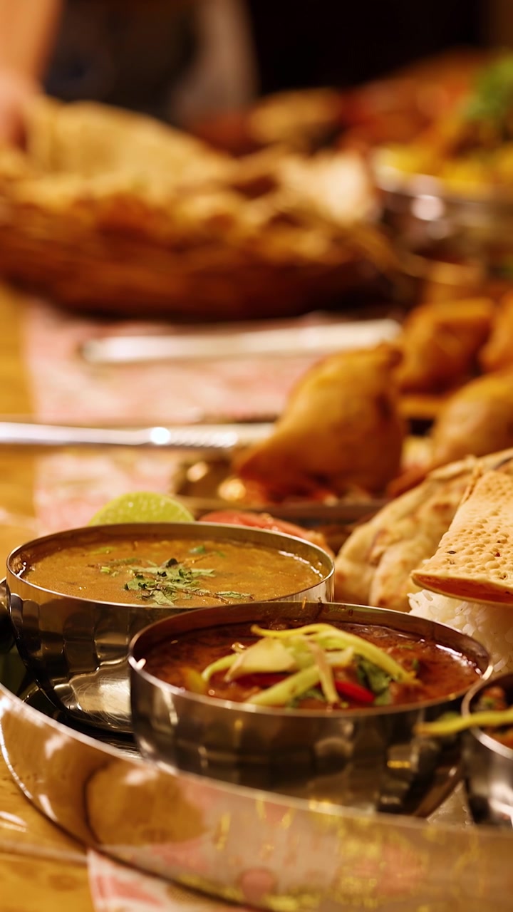 Close-up tracking shot of traditional Indian thali featuring various curries, samosas, and flatbreads.