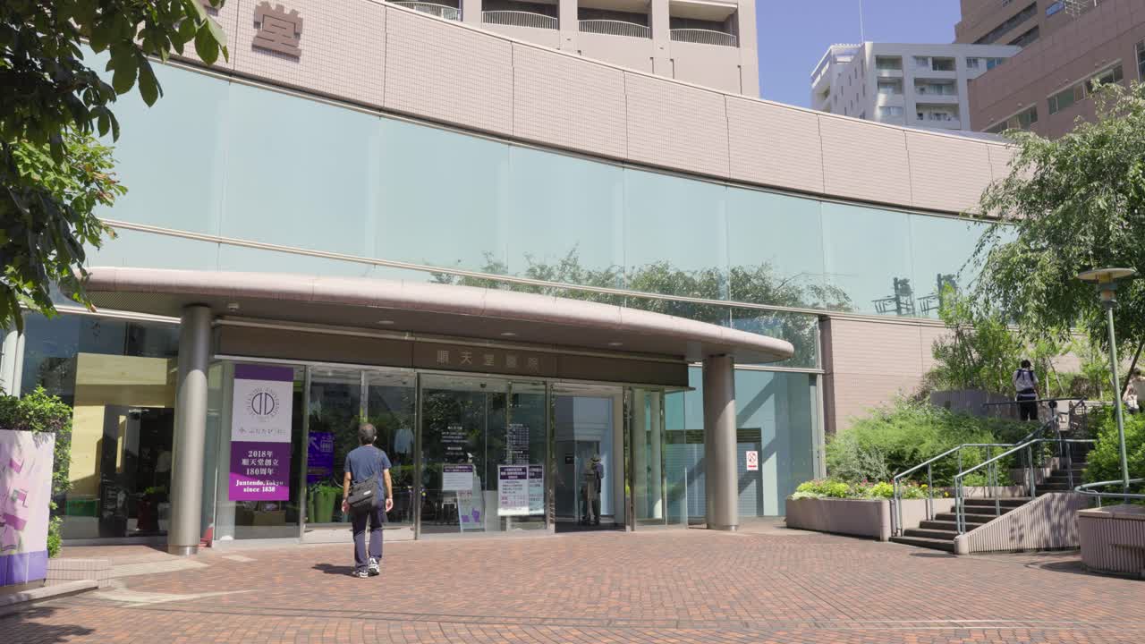 People entering and exiting a modern Tokyo hospital on a sunny day, surrounded by greenery