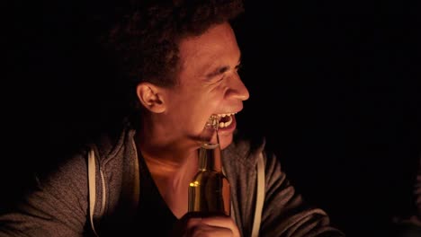 Attractive-african-american-opening-a-beer-bottle-with-teeth-and-giving-it-to-his-girlfriend-while-sitting-by-the-bonfire-late