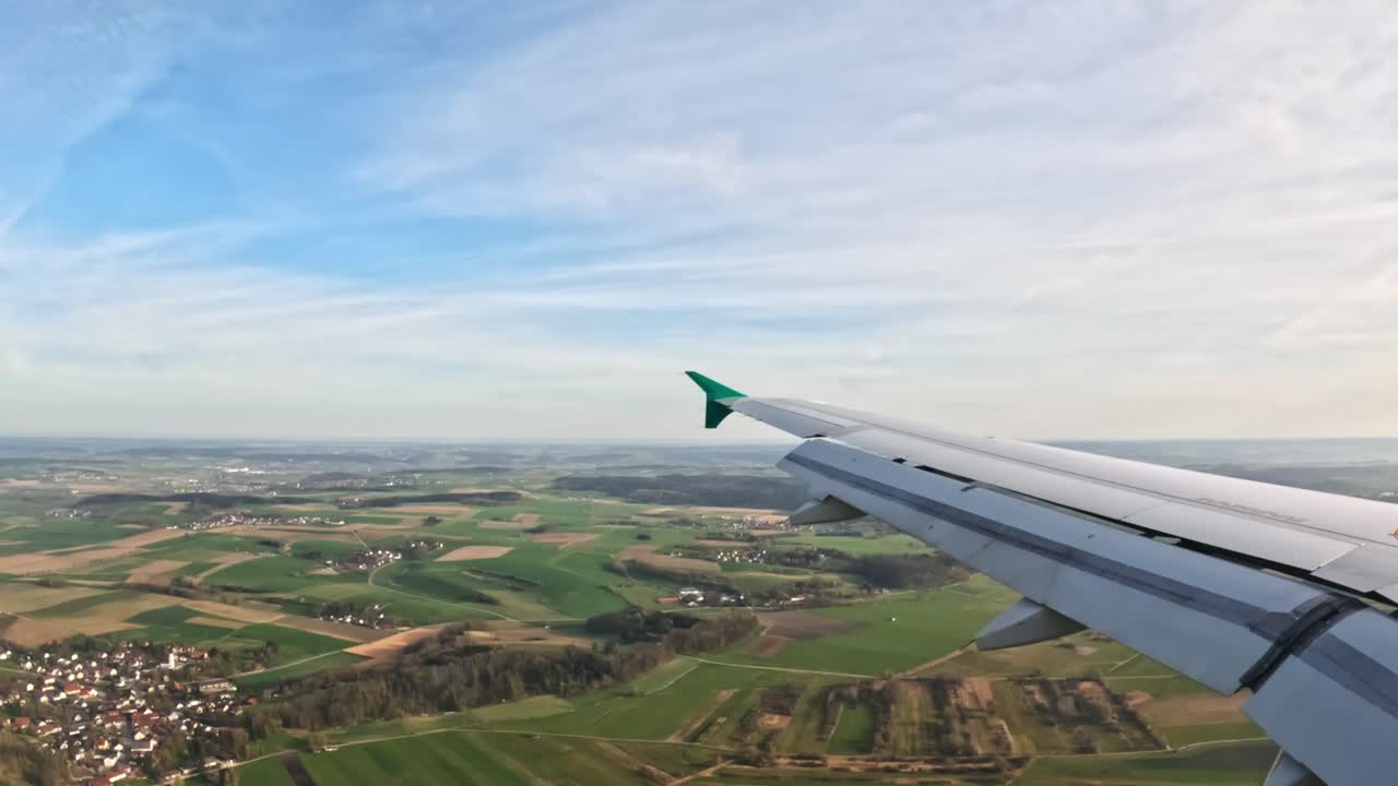 Capturing a stunning aerial view of the European countryside during a flight, showcasing vast fields, small towns, and a clear sky. The airplane wing adds a dynamic perspective to the scene
