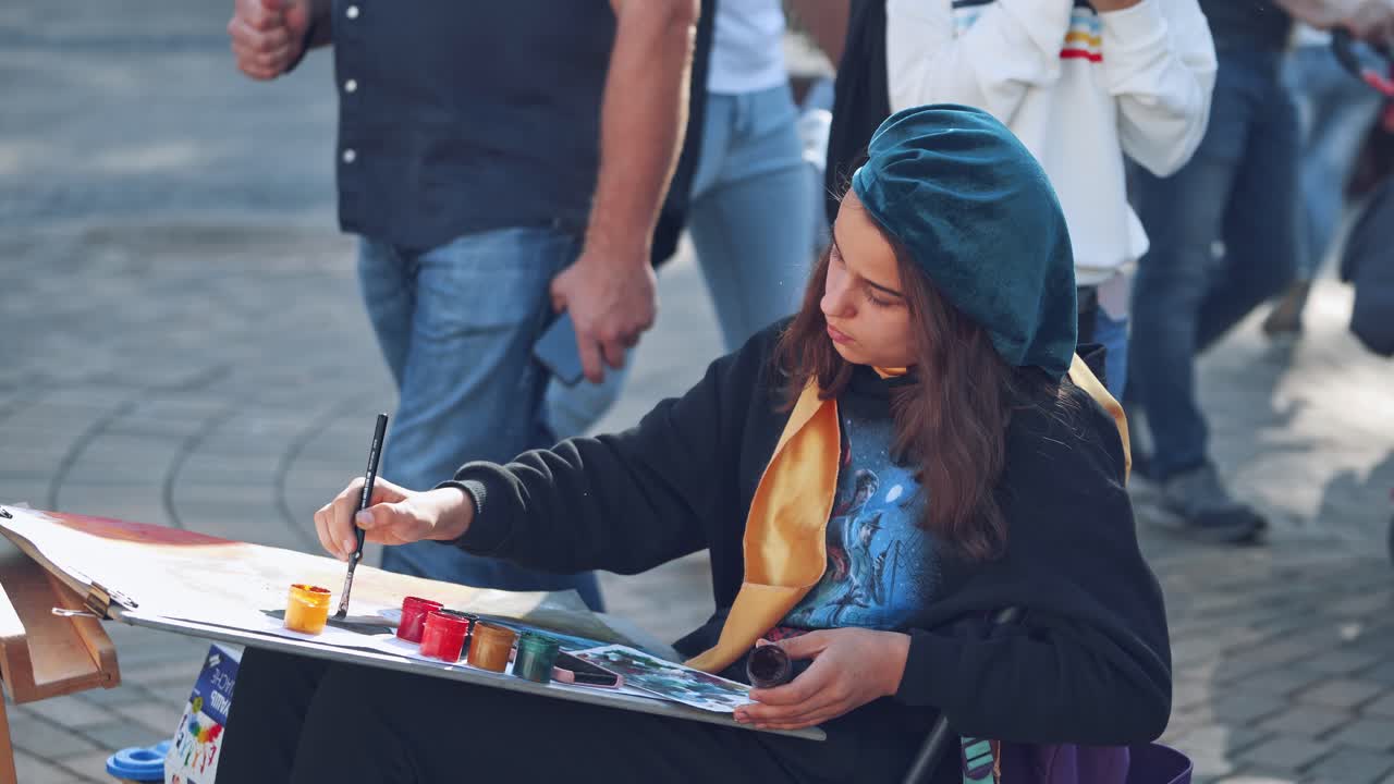 Female artist painting in the city. Young student paints picture with brush and colors on the busy street background.