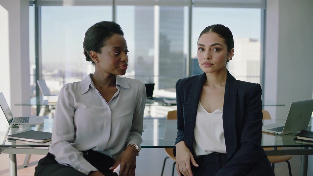 Turning left woman initiating update as peer listening in office attire at glass desk with laptops
