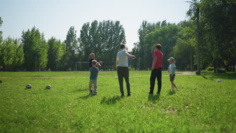 a grandfather leads his two grandsons in stretching their arms on a grassy field, while two younger children stand by with a soccer ball in the background