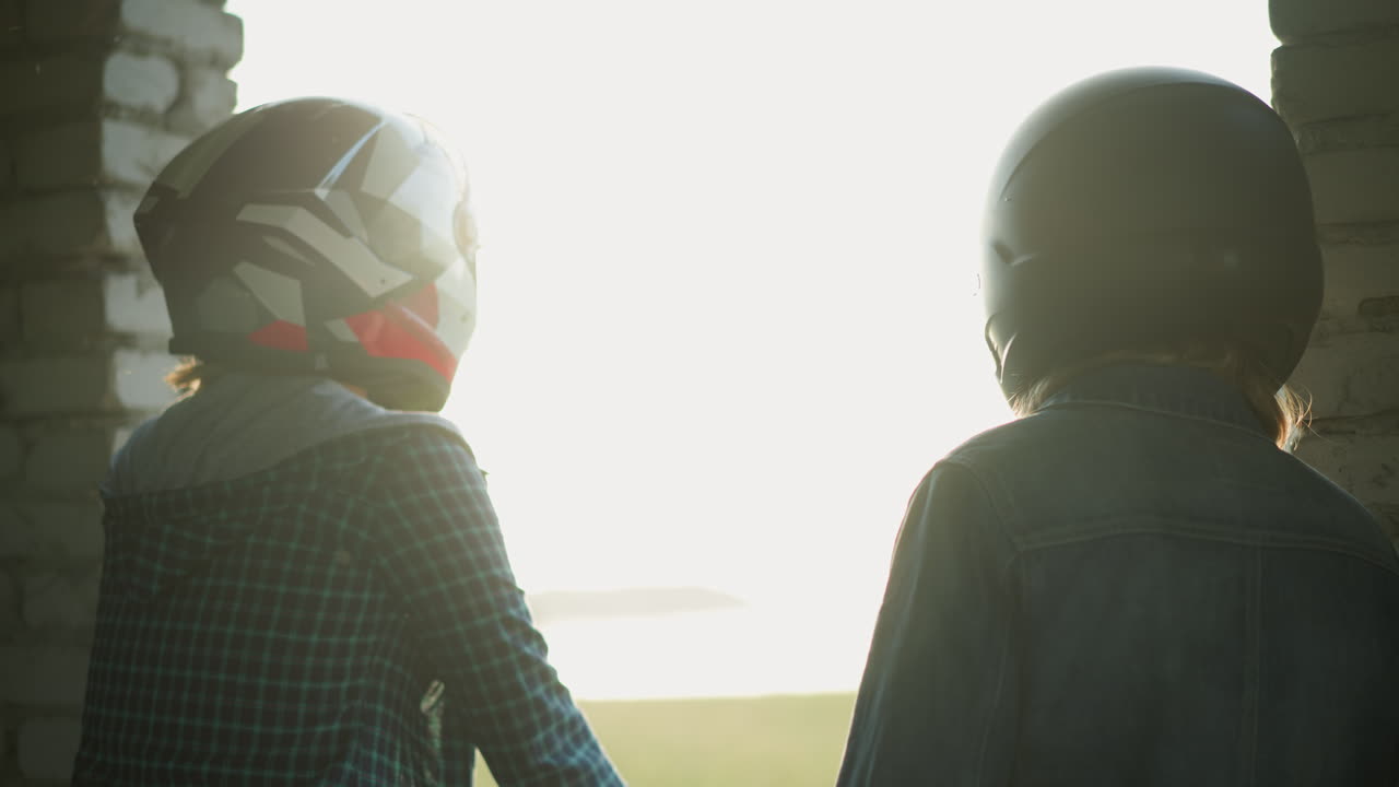 Back view of two women in helmets standing close to a window, looking outward toward a blurred structure in the background