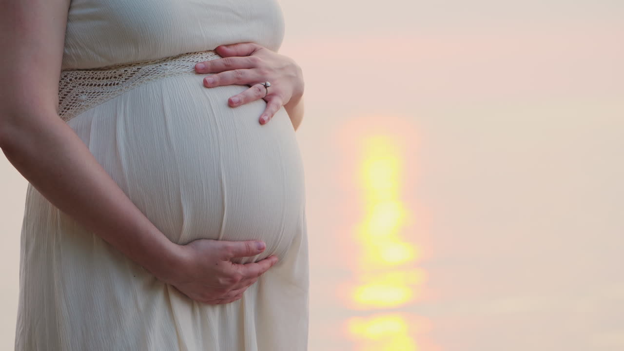 임신 한 여자는 그녀의 배를 쓰다듬어 그녀의 침대 클로즈업의 뒤에 바다 근처에 서있다