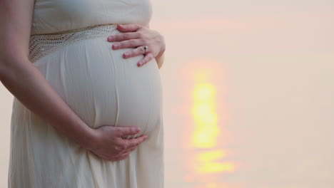 a pregnant woman is standing near the sea at the back of her bed close-up stroking her belly