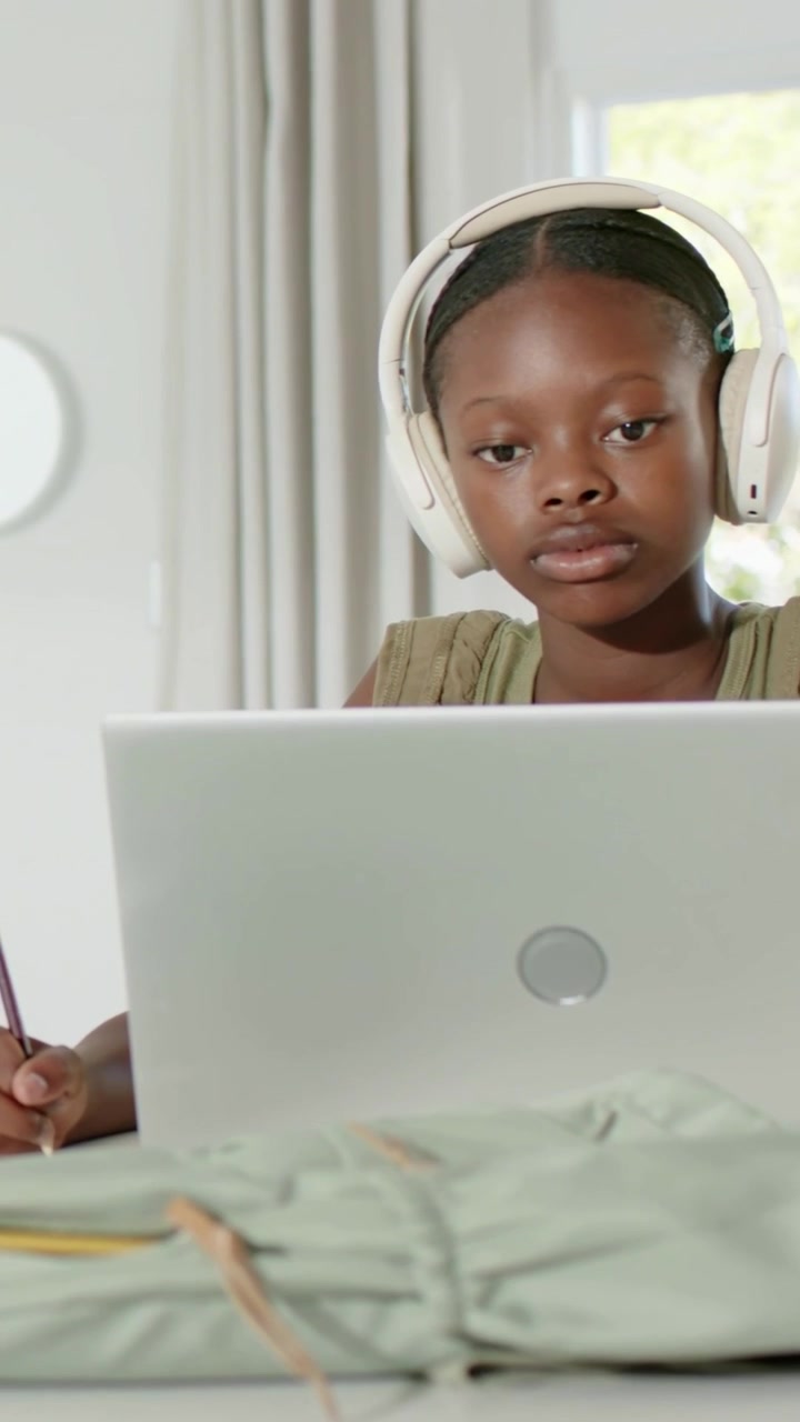 Vertical video: Seeing laptop, teen wearing olive top noting with pencil at desk, white headphones