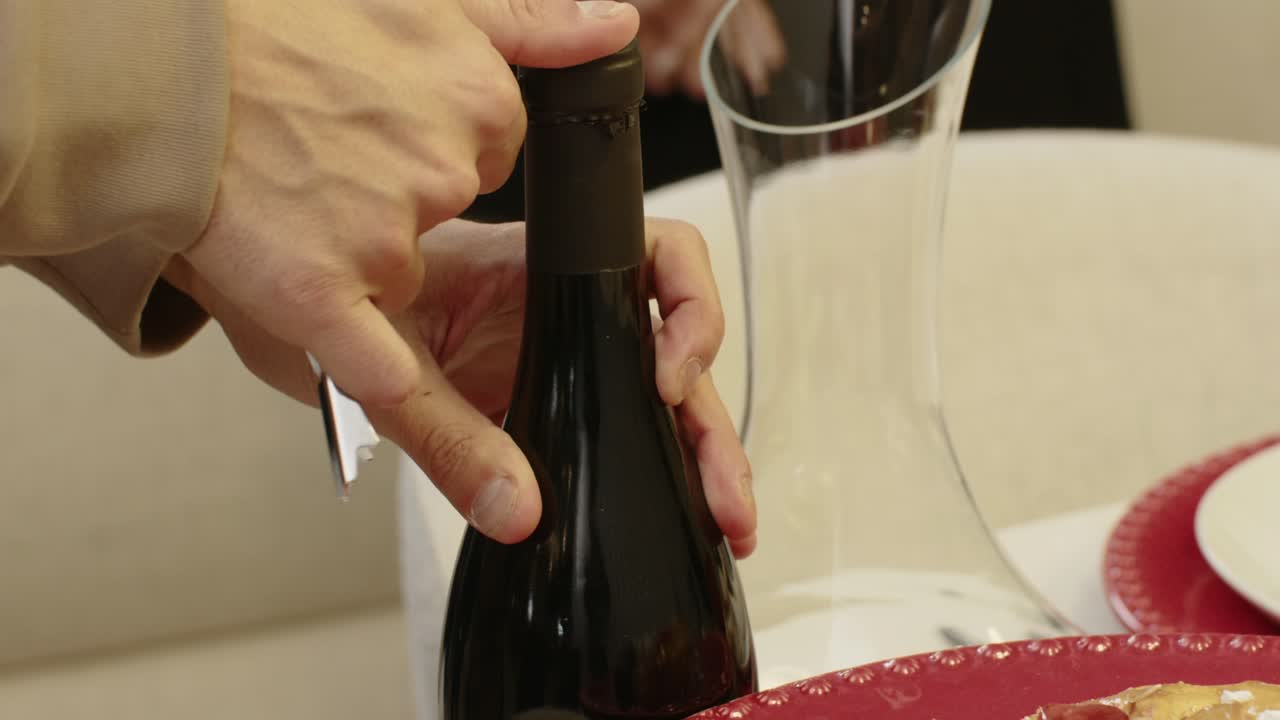 Close up of hands opening a wine bottle beside a glass decanter on a dining table