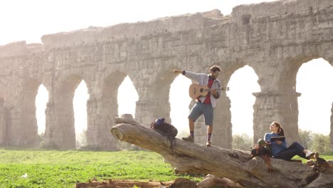 happy young couple backpackers tourists on a log trunk pointing direction with hand in front of ancient roman aqueduct ruins in parco degli acquedotti park in rome at sunrise romantic slow motion