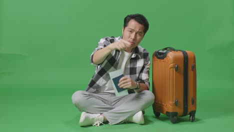 full body of asian male traveler with luggage and passport looking at camera and shouting at someone while sitting in the green screen background studio