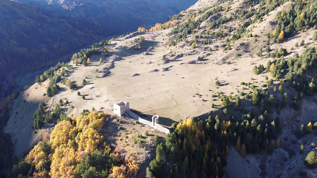 Drone captures a wide alpine valley with historic stone ruins surrounded by autumn forests. Rugged mountain peaks and clear November sunlight highlight the dramatic landscape in the Hautes Alpes