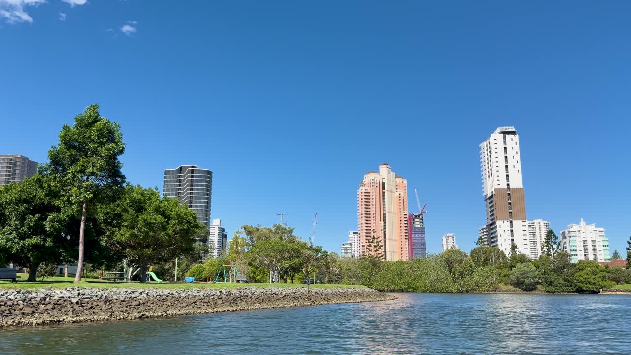 Camera glides over river toward modern skyscrapers, green park, and convention center under clear sky