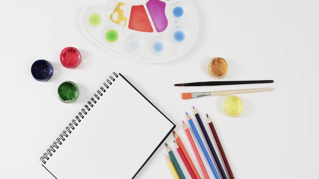 vista aérea del cuaderno de notas, pinturas, pinceles y lápices de colores sobre fondo blanco, en cámara lenta