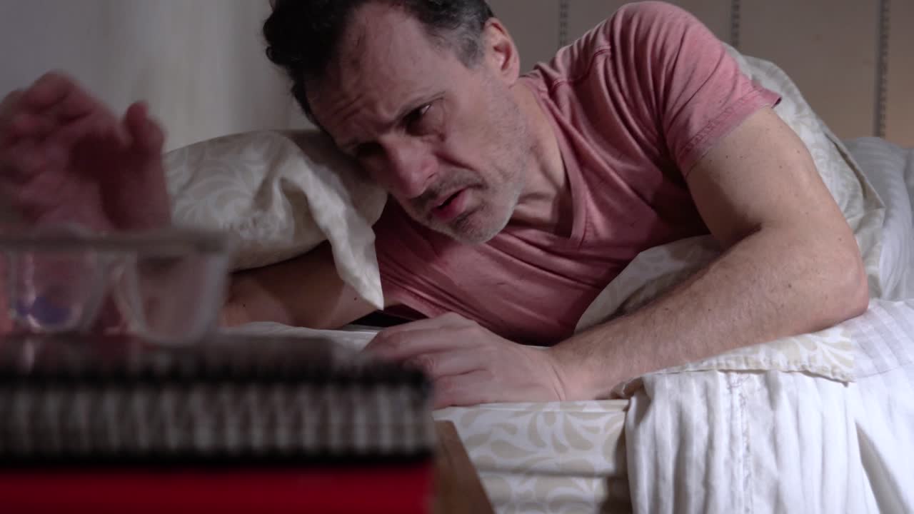 라틴계 남성이 아침에 전화를 확인하고 침대에 물 한 잔과 함께 약을 먹습니다.