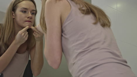 young woman squeezes pimples on her chin while standing in the bathroom in front of the mirror