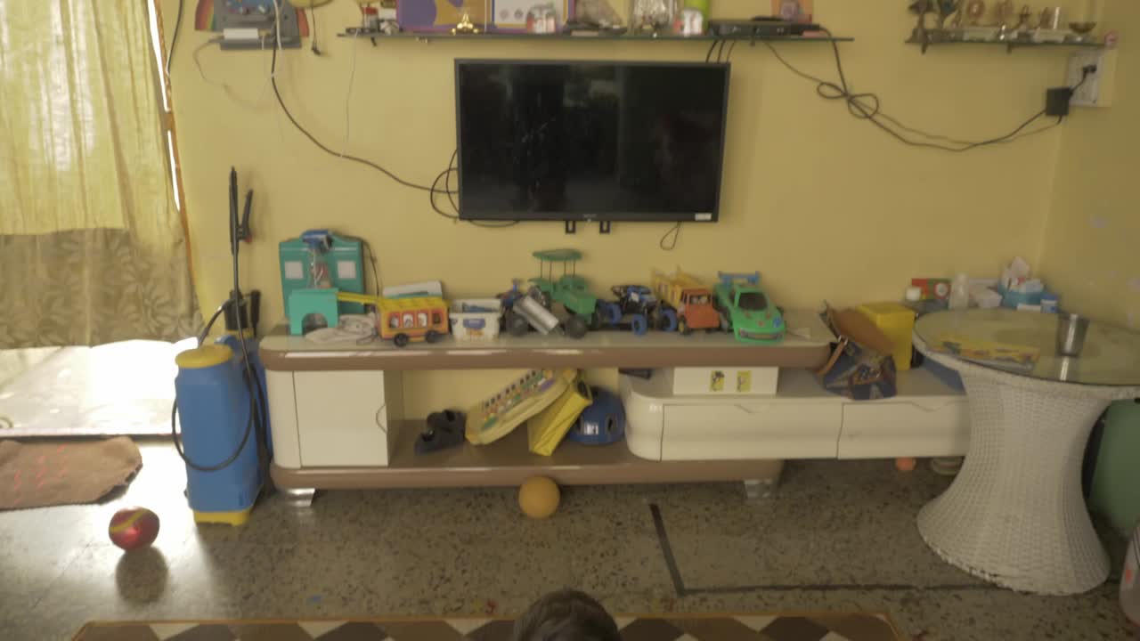 양탄자에 누워 홀에서 혼자 텔레비전을 보고 있는 아시아계 어린 아이와 장난감과 트로피로 장식된 텔레비전 유닛과 캐비닛을 보여주는 틸트업 샷