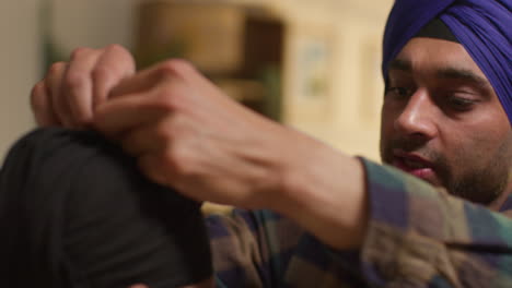 close up of father tying turban onto head of young sikh son with top knot sitting on sofa at home 1