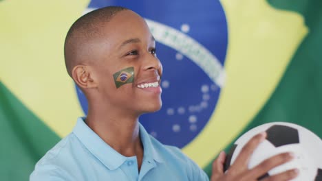 Video-De-Un-Niño-Afroamericano-Feliz-Sosteniendo-Una-Pelota-Sobre-La-Bandera-De-Brasil-Viendo-El-Partido